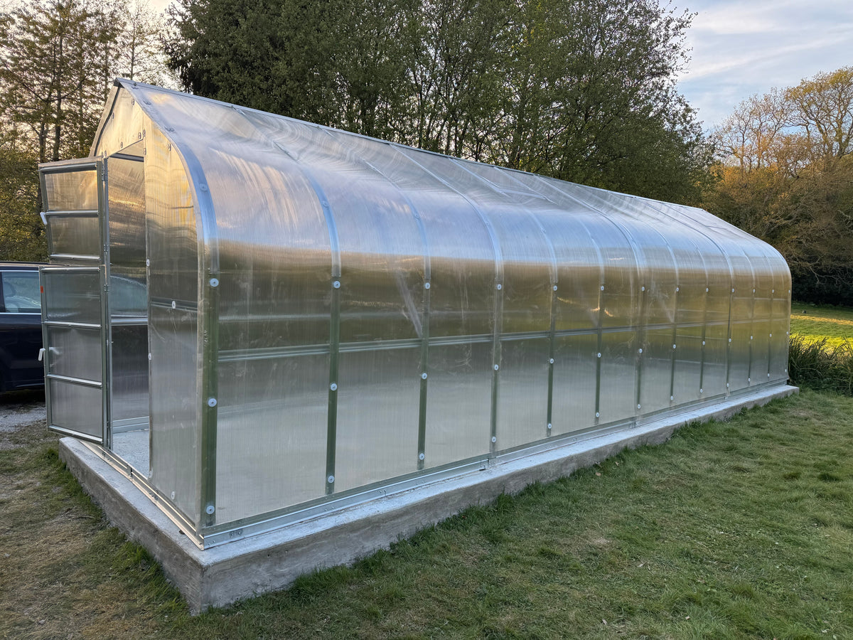 Polyeco Spade 8m long greenhouse with 6mm polycarbonate cover, mounted on a concrete base in an outdoor grassy area with trees in the background.