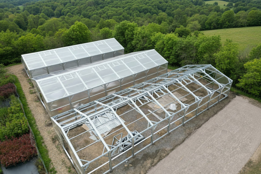 An aerial view shows two large greenhouse structures on a landscaped site. One greenhouse has a translucent white roof and a silver metal frame, while the other is mostly open with only a partial silver frame.