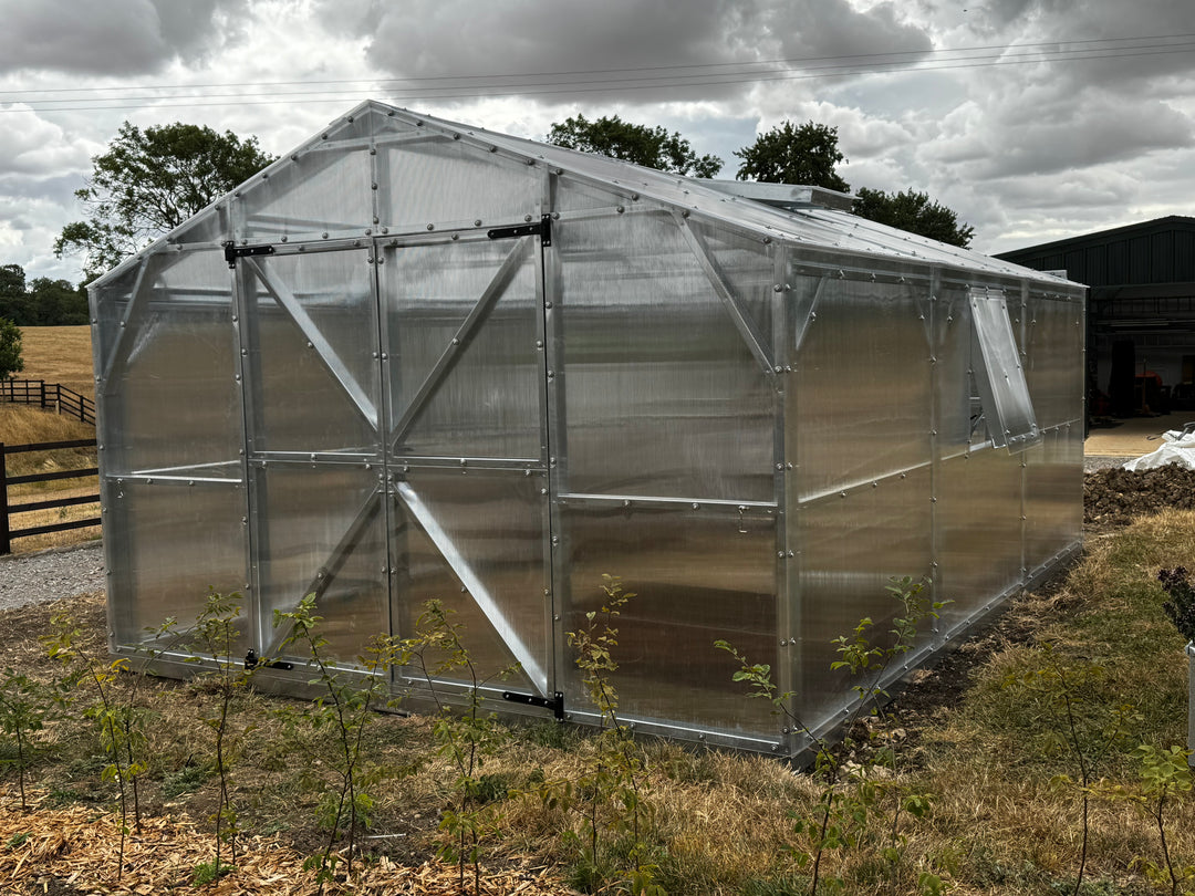 A large greenhouse with a silver metal frame and translucent polycarbonate panels, situated outdoors on grassy ground under a cloudy sky.