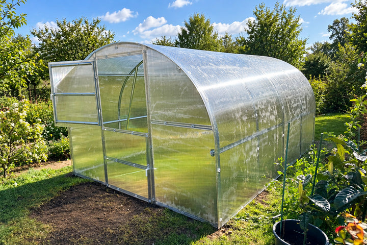 A small arched greenhouse with transparent plastic panels and a silver metal frame, set outdoors in a garden with grass, plants, trees, and a blue sky with clouds.