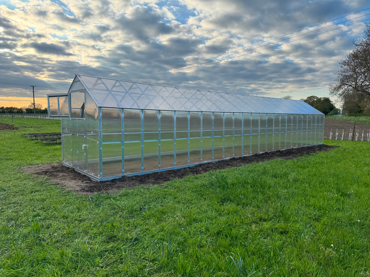 A long, low-profile greenhouse with a transparent polycarbonate or acrylic panel roof and frame, situated on a grassy field with a cloudy sky in the background.