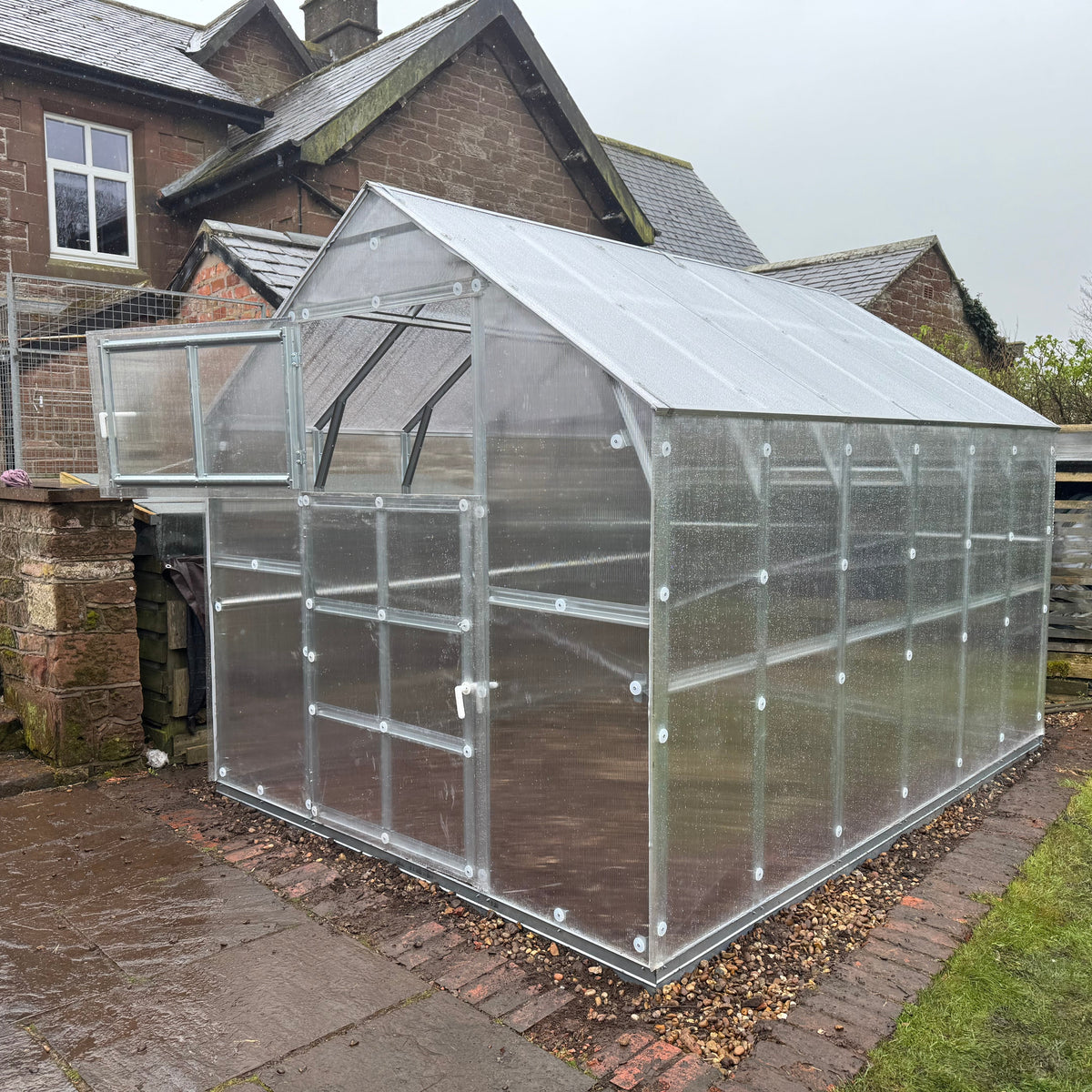 Polyeco House polycarbonate greenhouse, shown installed in a backyard next to a brick house.