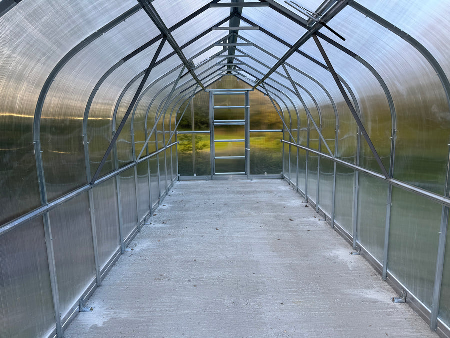 A long walkway greenhouse or hoop house with a concrete floor, clear translucent side panels, arched metal frame structure, and a glass door at the far end.