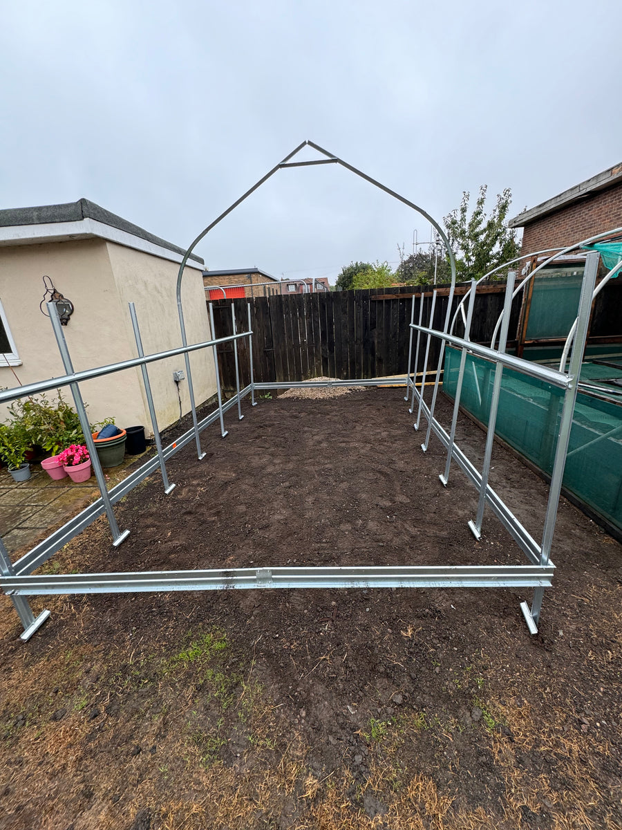 A metal garden hoop house frame with a rectangular base and arched roof support, set up on a patch of bare soil in a backyard with a fence, house wall, and green tarp visible.
