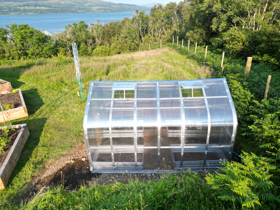 A small arched polytunnel greenhouse with a silver aluminum frame and clear plastic panels, situated on a grassy hillside with a lake and forest in the background.
