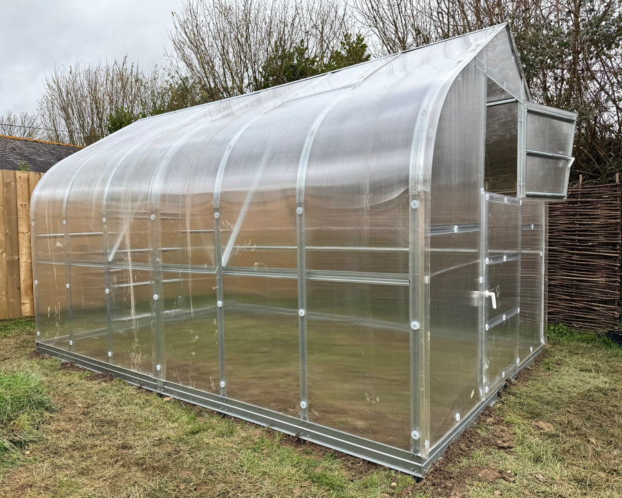 A small arched greenhouse with a transparent plastic cover and silver metal frame, shown outdoors on grass with trees and a wooden fence in the background.