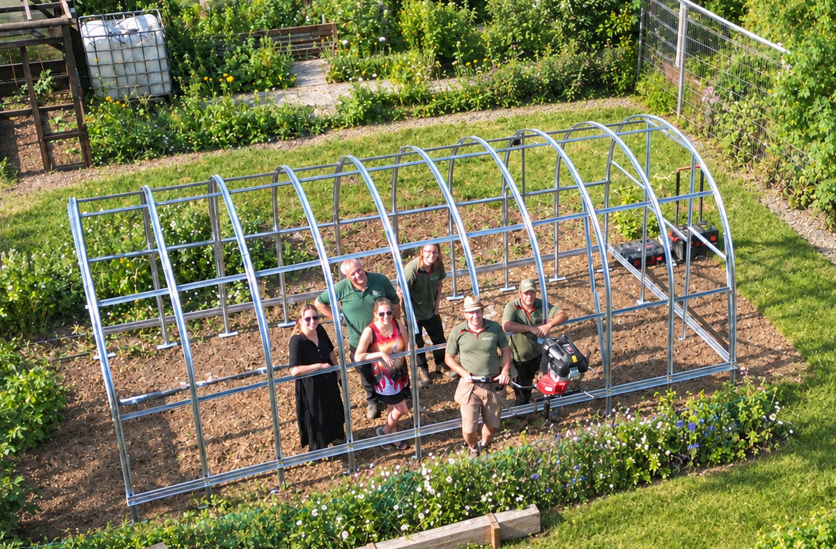 A group of people standing and sitting under a large galvanized steel greenhouse frame in a backyard garden setting with plants, containers, and other garden equipment visible.